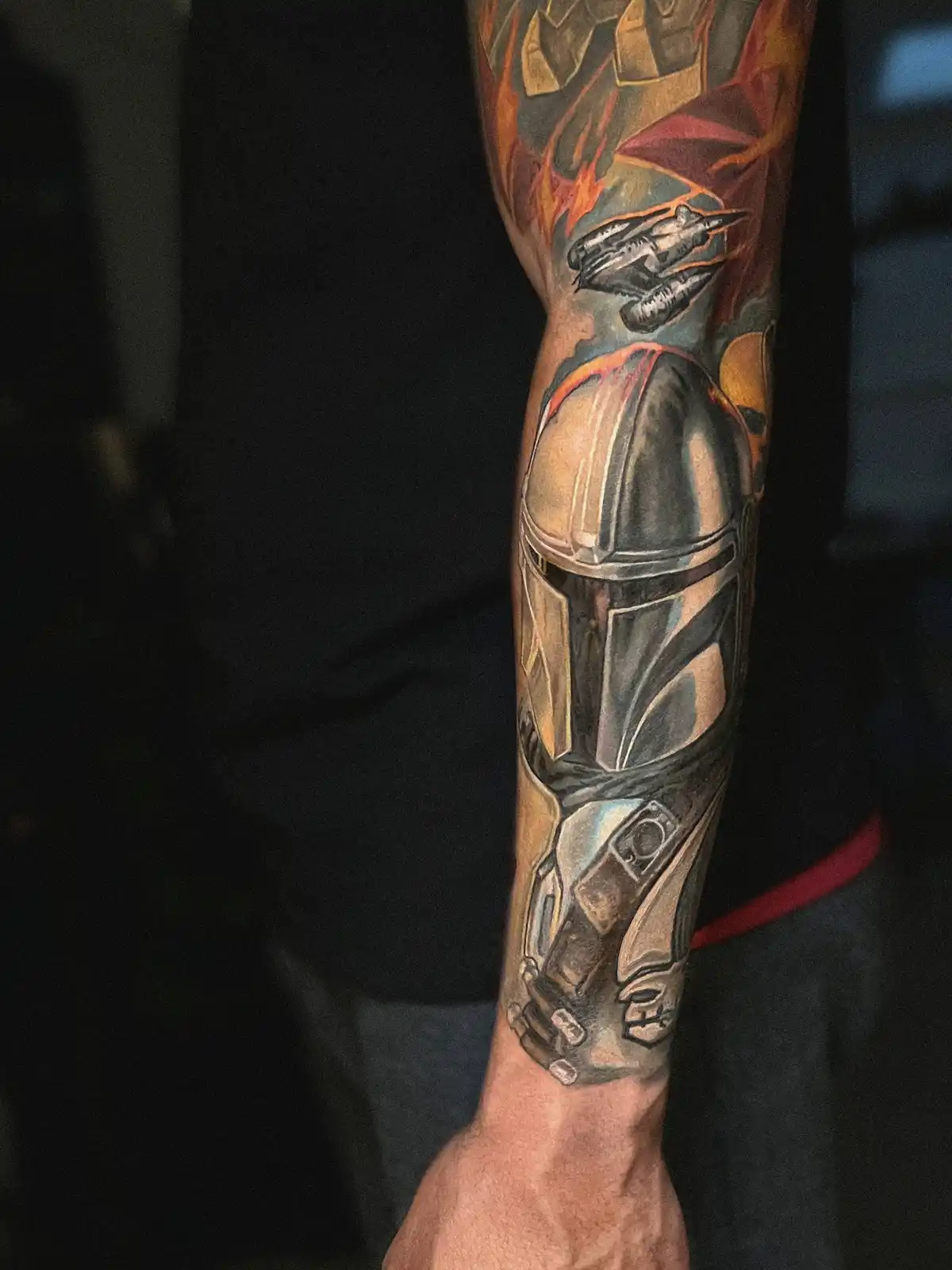 A person's forearm is covered in a detailed tattoo depicting the Mandalorian helmet, a spaceship, and flames. The overlaid…