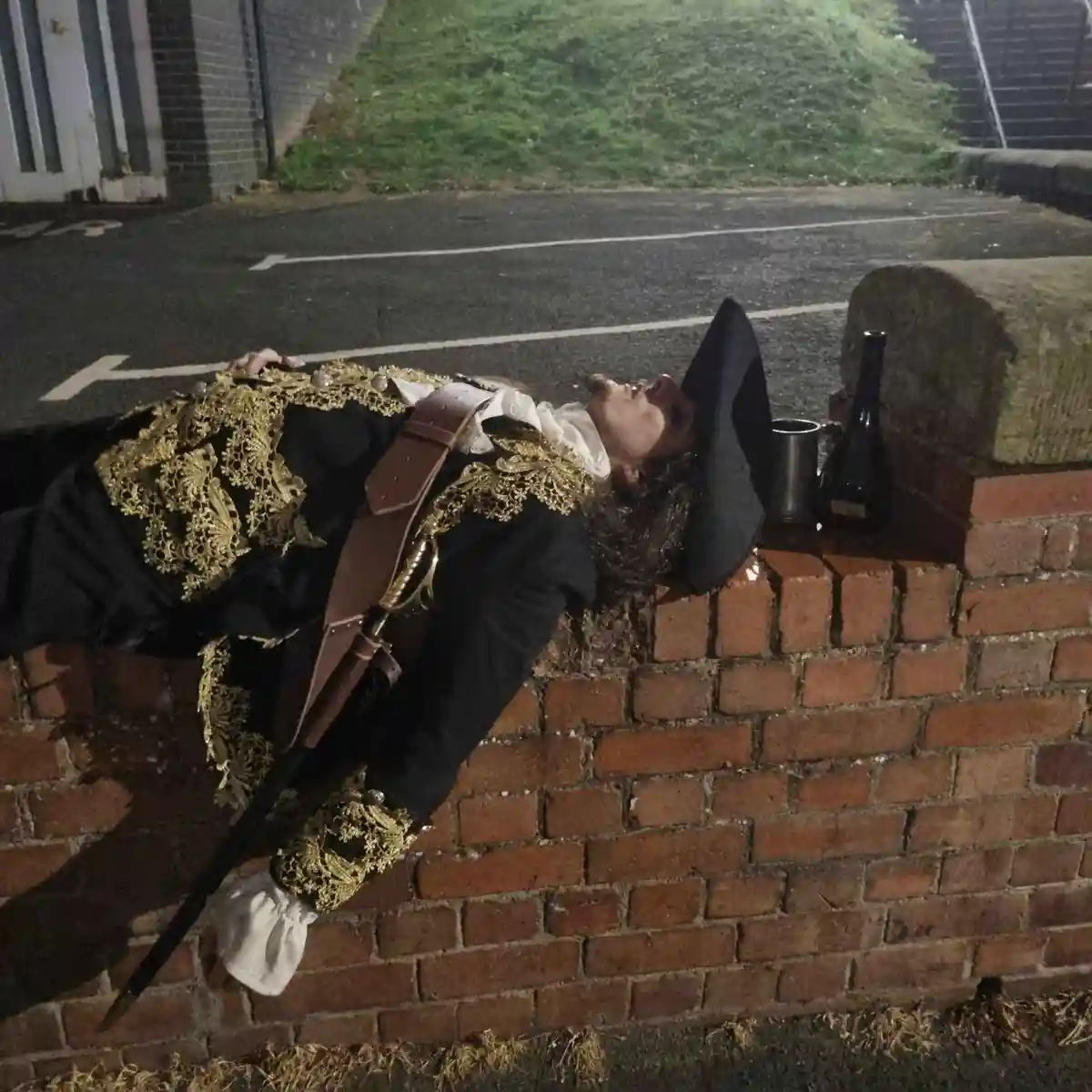 A person dressed in ornate pirate costume, including a tricorn hat and gold-embroidered jacket, lies passed out on a low…