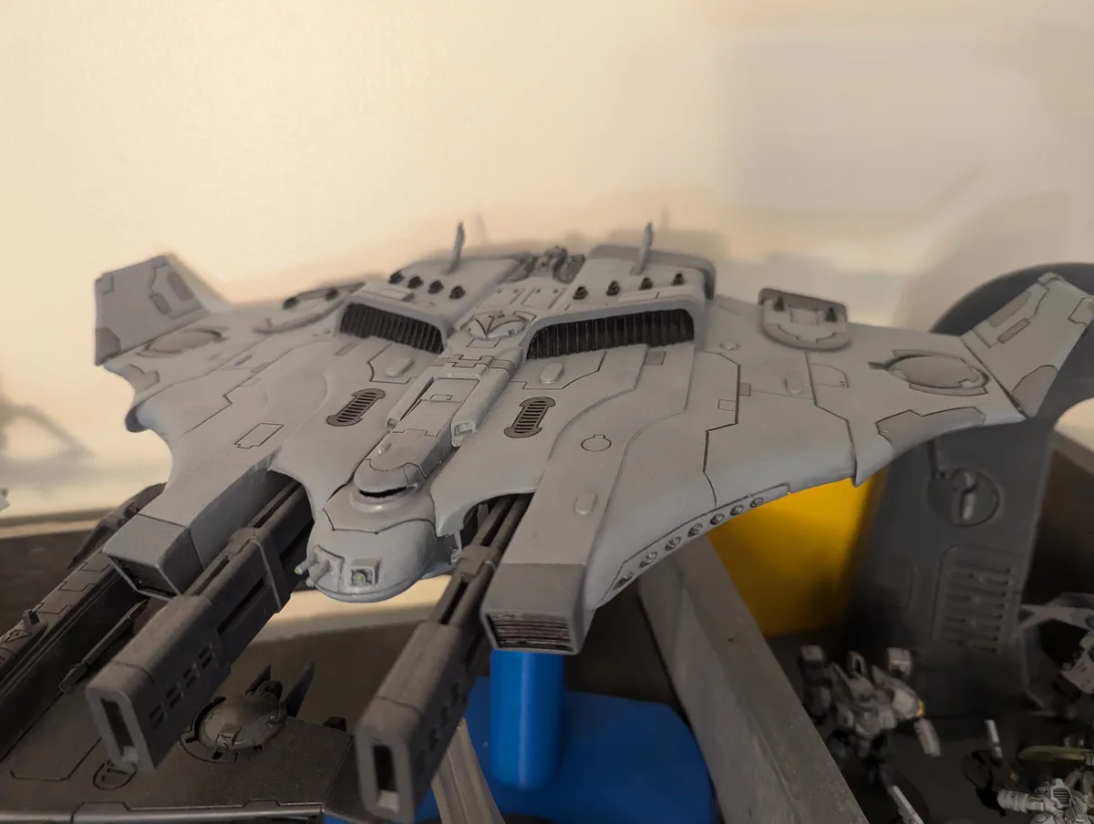 A gray, unpainted plastic spaceship model, likely from a tabletop wargame, is displayed on a blue stand. The model has…