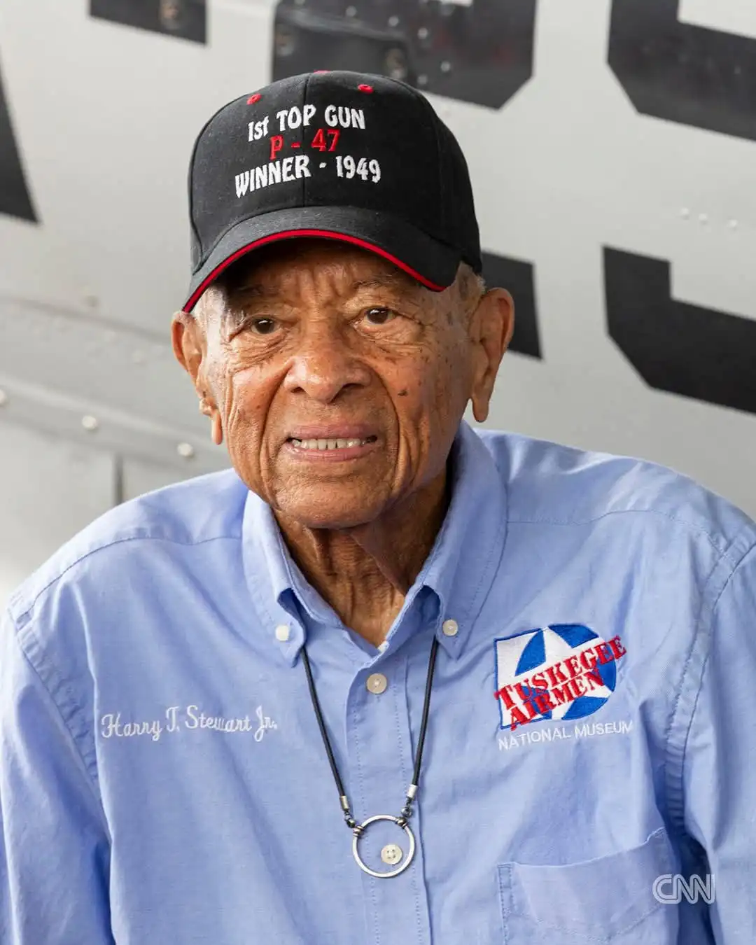 An elderly Black man, identified by his shirt as Harry T. Stewart Jr., wears a black cap that reads '1st TOP GUN P-47 WINNER…
