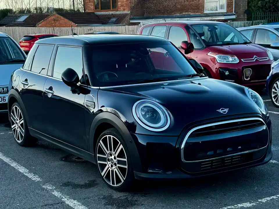 A black Mini Cooper electric car is parked in a lot with other vehicles, including a red Citroën. The car has distinctive…