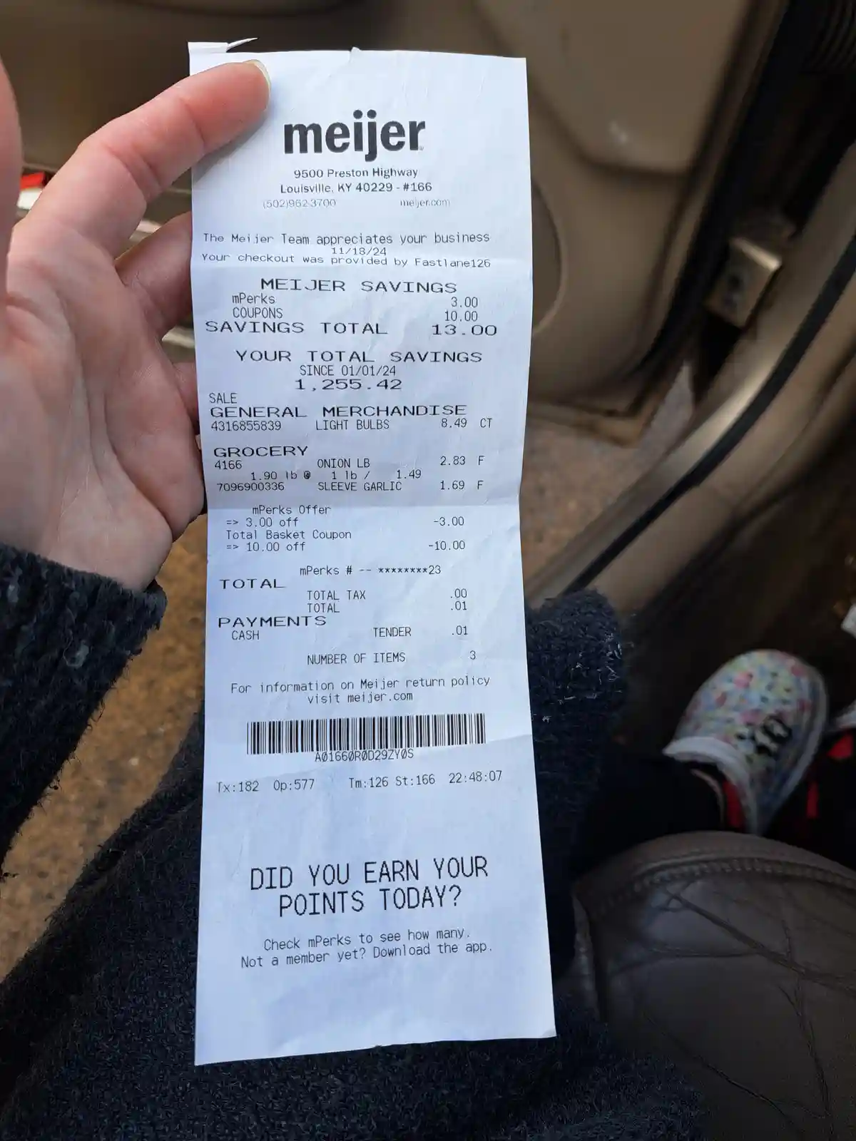 A person holds a Meijer grocery receipt inside a car, showing a total of $0.01 after $13.00 in savings. The receipt's bottom…