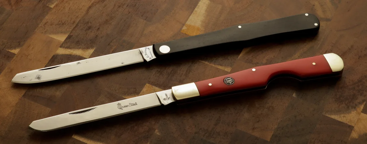 Two steak knives lie on a wooden cutting board; one has a black handle and the other a red handle, both with visible brand…