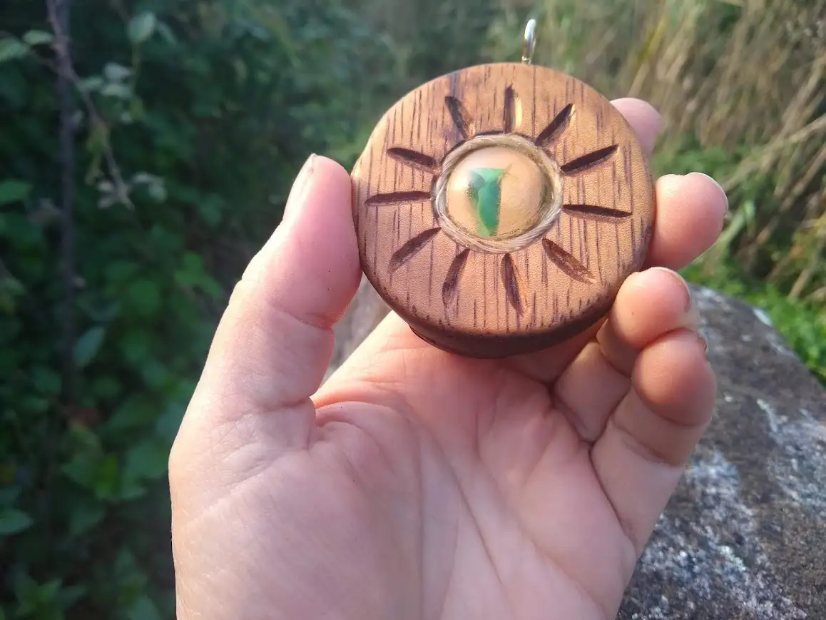 A hand holds a small, round wooden pendant carved with a sun design, featuring a clear circular center containing a green…