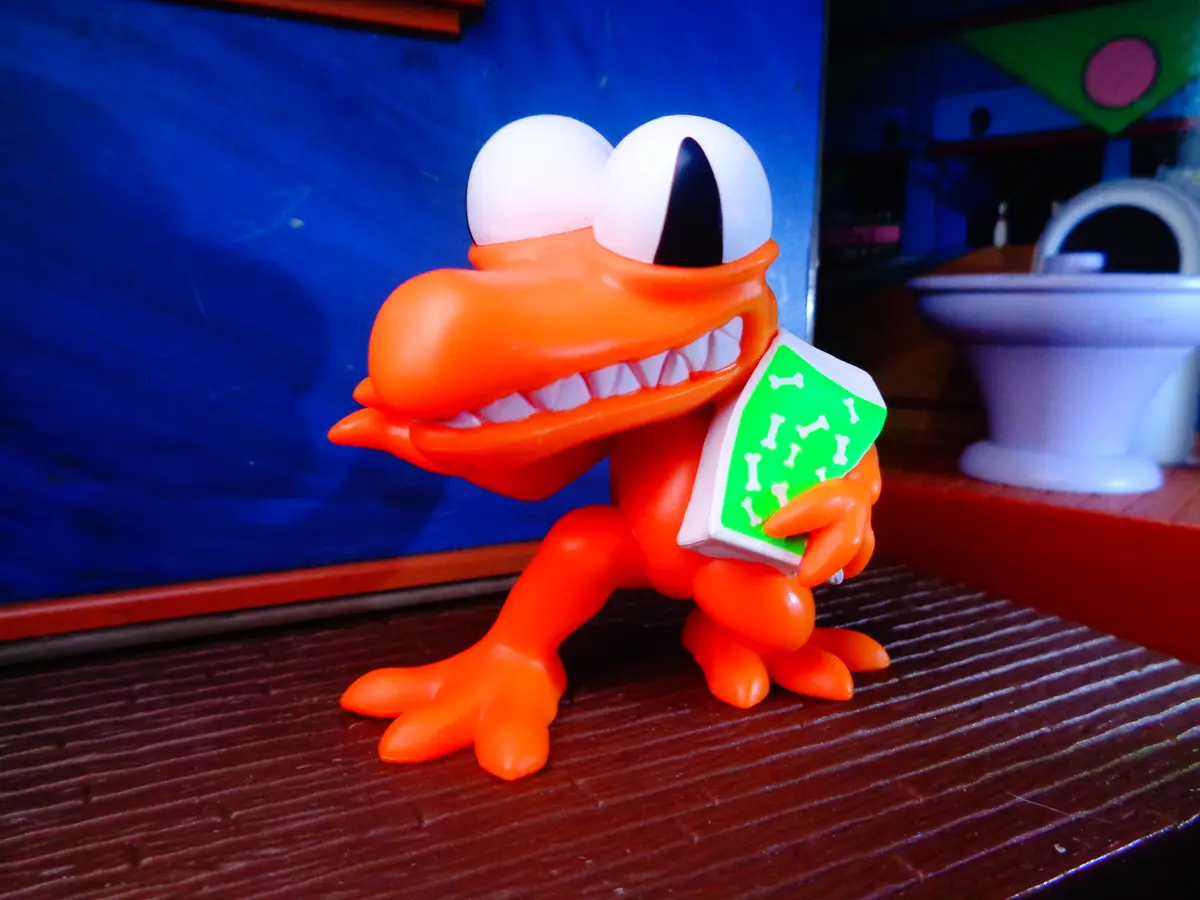 An orange plastic toy crocodile with large white eyes and a wide grin stands on a wooden surface, holding a green box…