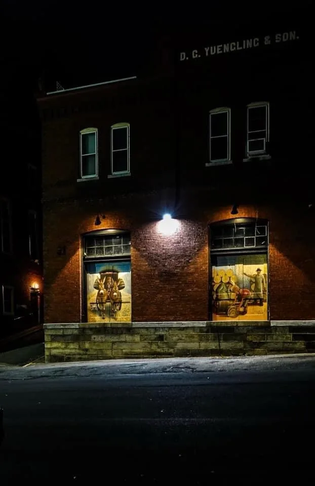 A nighttime photo of a brick building with 'D. G. YUENCLING & SON.' written on the roof. Two large windows display…