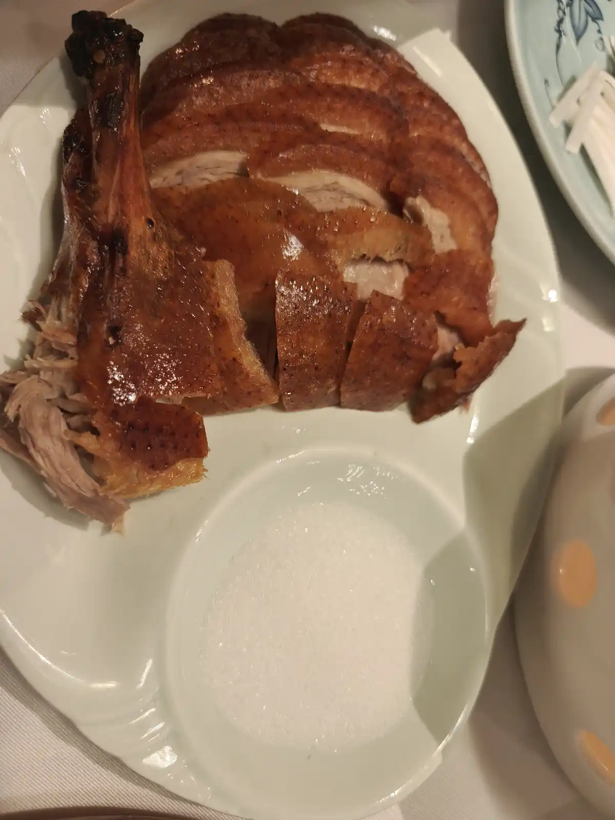 A plate of sliced roasted duck with a glossy, caramelized skin sits next to a small dish filled with granulated sugar…
