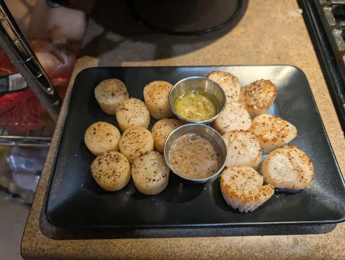 A black rectangular plate holds two groups of seared scallops, one group with a golden-brown sear and the other with a…