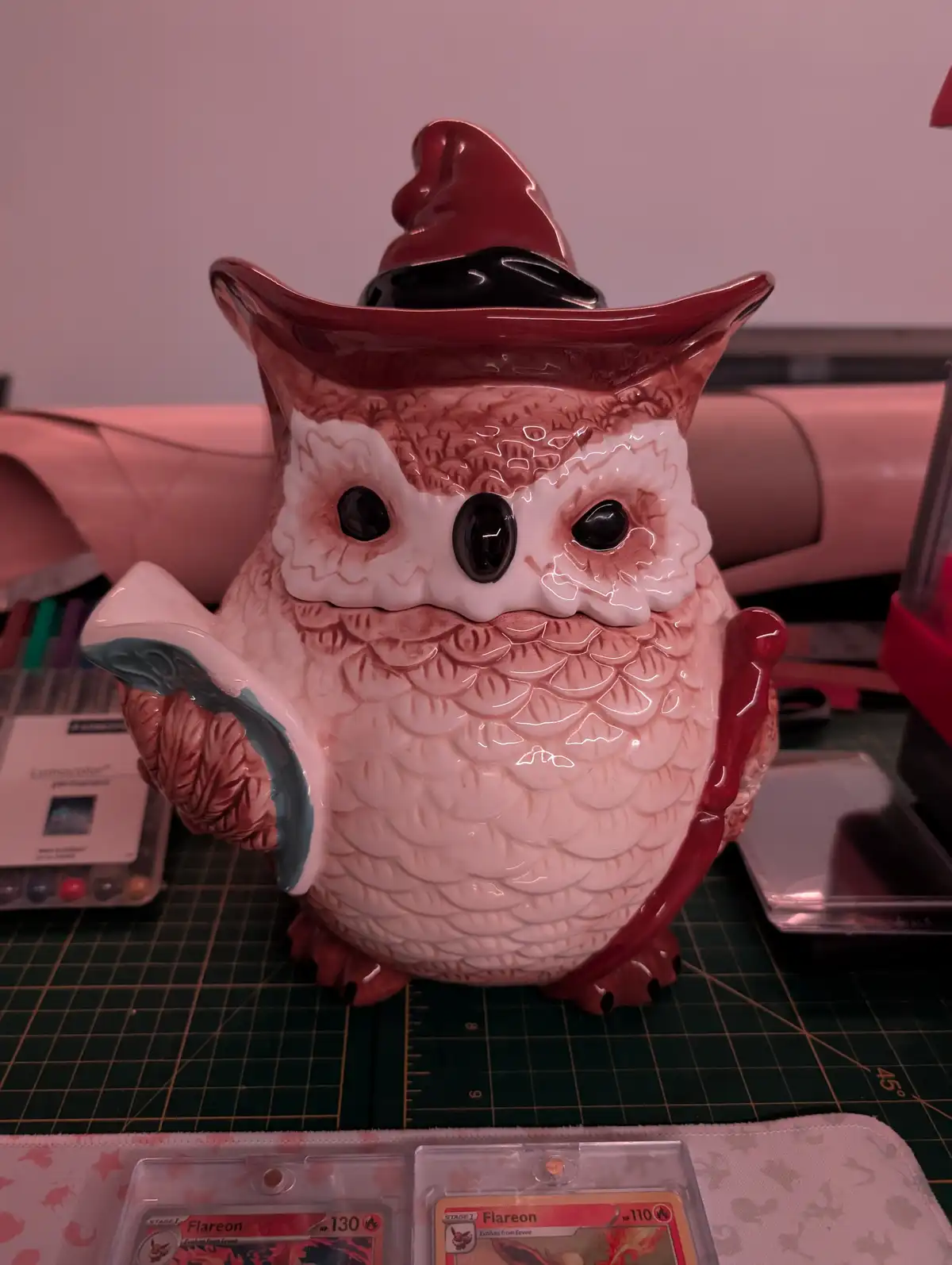 A ceramic owl-shaped teapot with a red witch's hat and a red handle, sitting on a green cutting mat. Two Pokémon trading…