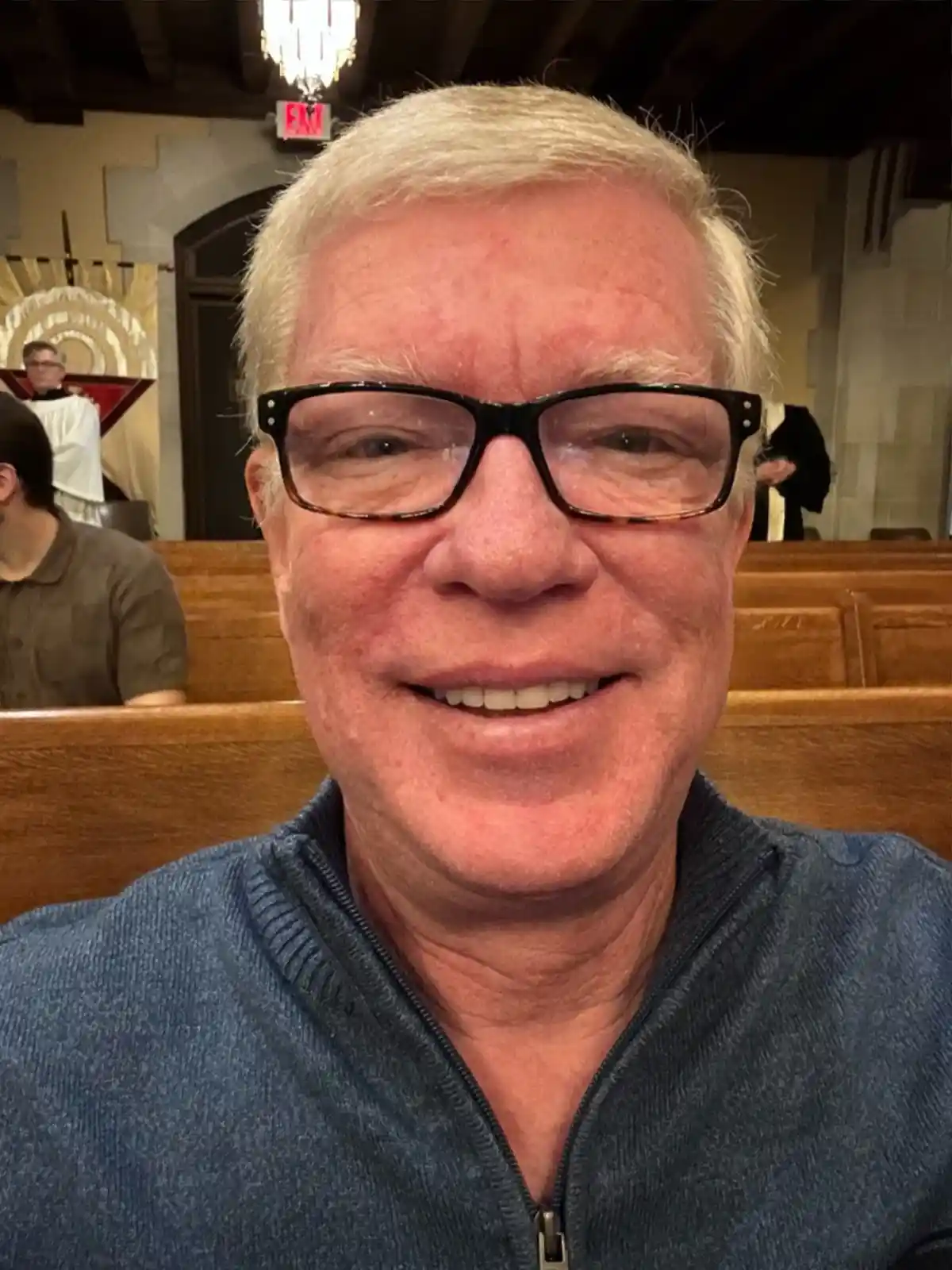 A close-up selfie of a smiling older man with white hair and black-rimmed glasses, wearing a blue zip-neck sweater, seated…