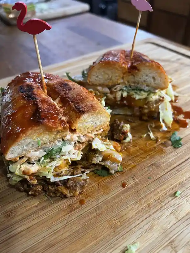 A large, messy sandwich cut in half sits on a wooden cutting board, with sauce dripping out and ingredients spilling over…