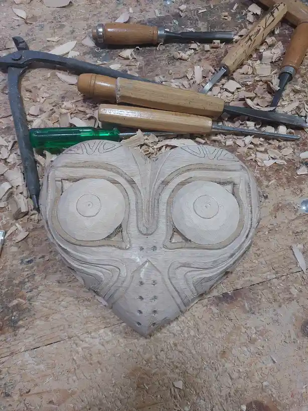 A partially carved wooden mask resembling Link's 'Mask of Truth' from The Legend of Zelda, surrounded by woodworking tools…
