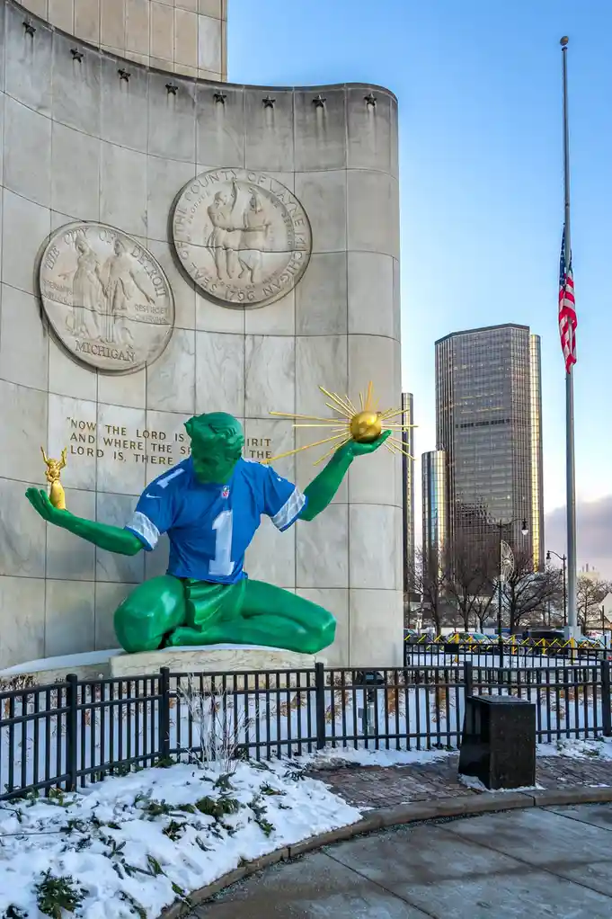 A green statue of a football player in a blue jersey with the number 1 sits cross-legged in front of a large stone monument…