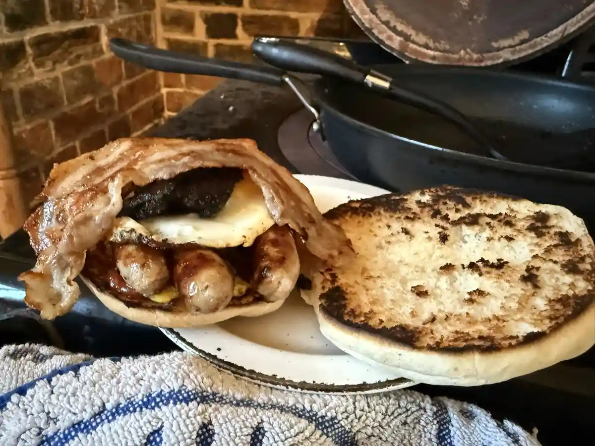 A close-up photo shows a toasted English muffin split open and stuffed with a fried egg, several sausages, a grilled patty…