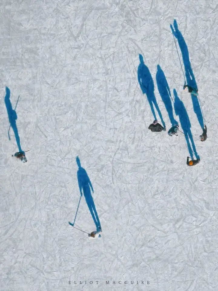 An aerial view shows several people ice skating on a frozen surface, their long blue shadows stretching across the textured…