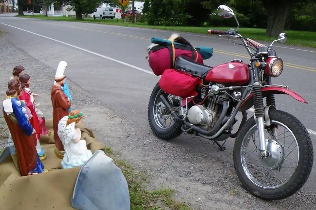 A red motorcycle with red saddlebags and a rolled-up sleeping bag strapped to the back is parked on a roadside gravel…