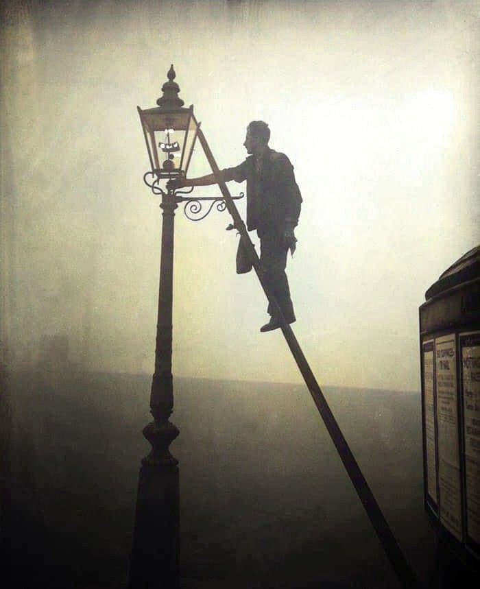 A vintage-style photograph shows a silhouetted man perched precariously on a tall, ornate lamppost, holding a ladder and…