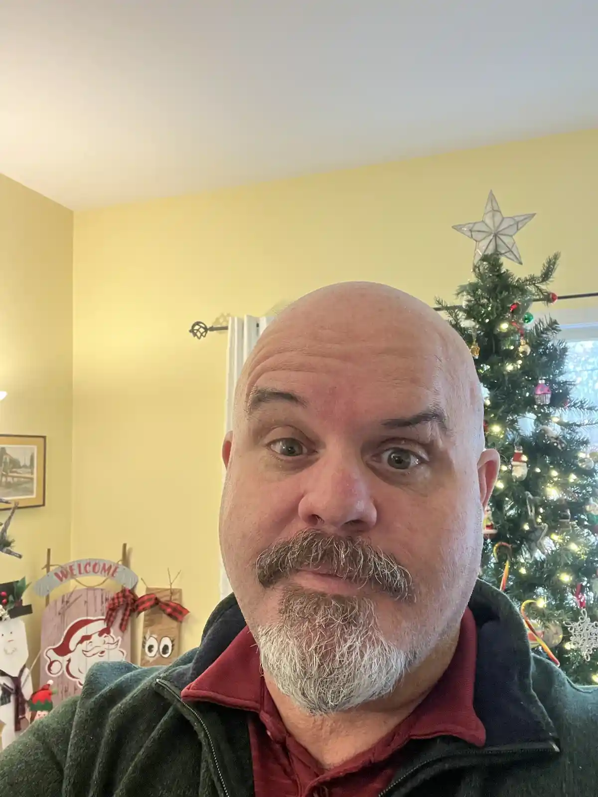 A bald man with a graying goatee and mustache stares directly at the camera with wide, surprised eyes. He wears a red…