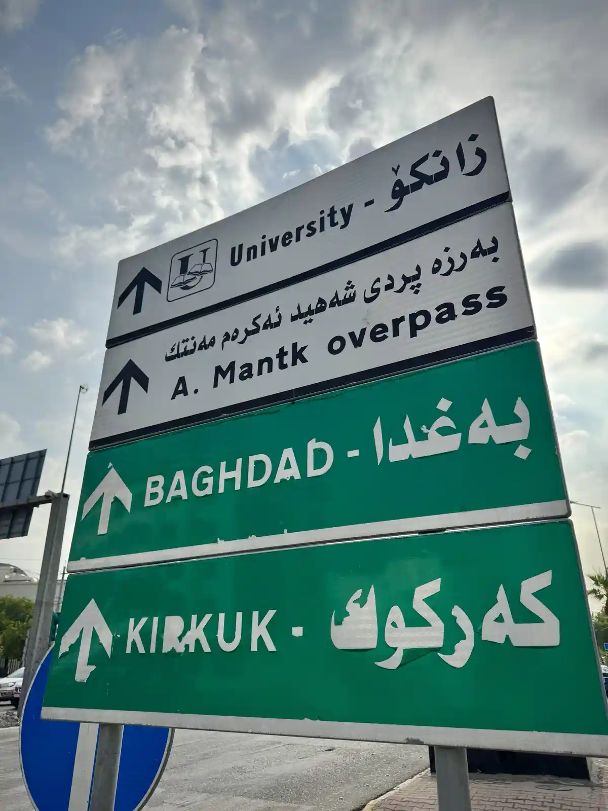 A multi-panel road sign with directional arrows pointing forward, featuring bilingual text in Arabic script and English. The…