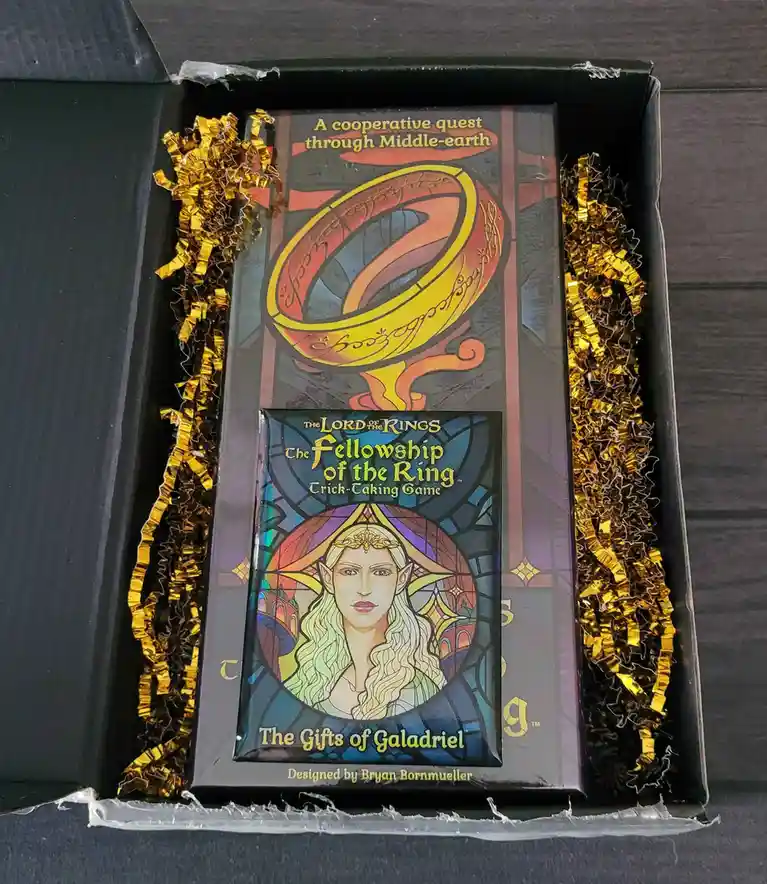 A top-down photo shows a black game box with gold crinkle paper on both sides, containing two Lord of the Rings card games…