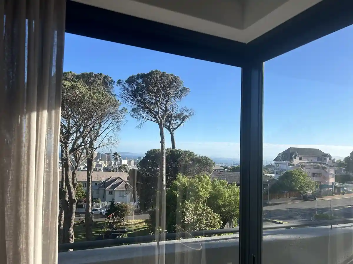 A photo taken from inside a room looking out through a large window at a sunny suburban scene with trees, houses, and a…