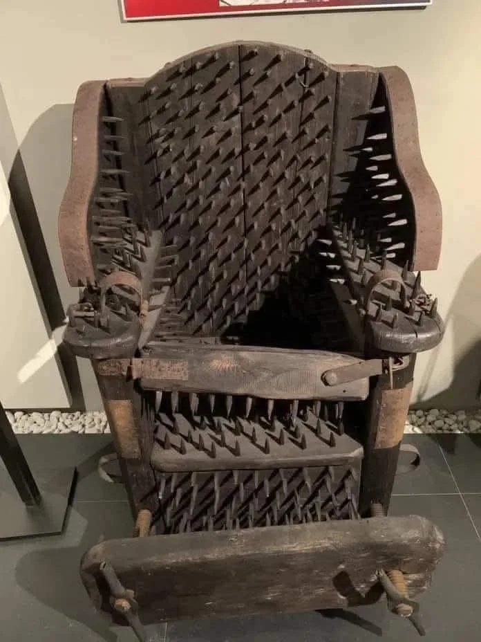 a dark, wooden torture chair covered in numerous sharp spikes protrudes from its back, arms, and seat, displayed in a museum…