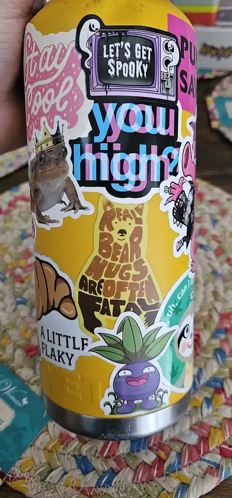 A yellow beverage can covered in colorful, overlapping stickers featuring cartoon frogs, bears, plants, and text like 'LET'S…
