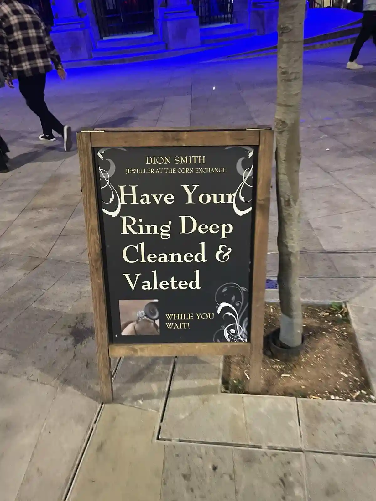 A wooden-framed sidewalk sign for a jeweler named Dion Smith advertises 'Have Your Ring Deep Cleaned & Valeted WHILE YOU…