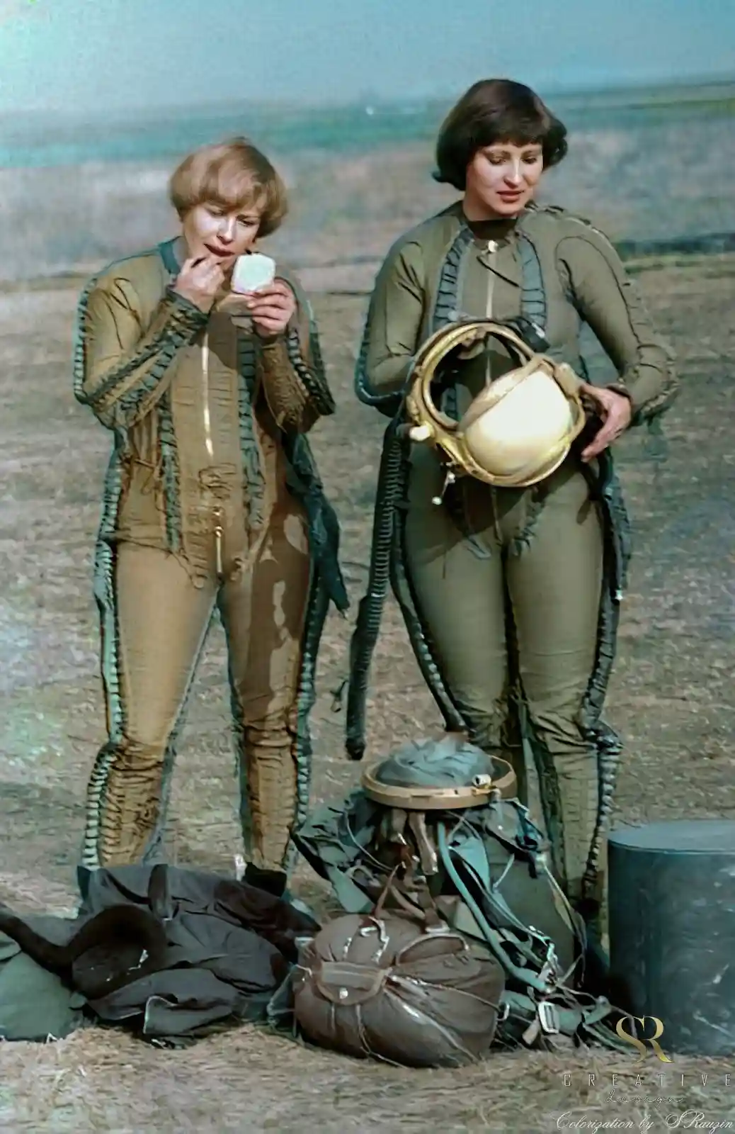 Two women in vintage-style flight suits stand outdoors, one applying makeup from a compact while the other holds a helmet…