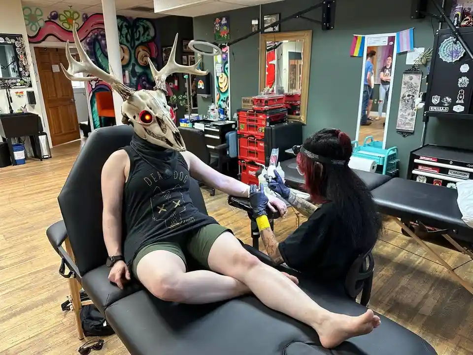 A person wearing a deer skull mask with glowing red eyes sits in a tattoo chair while a tattoo artist works on their leg…