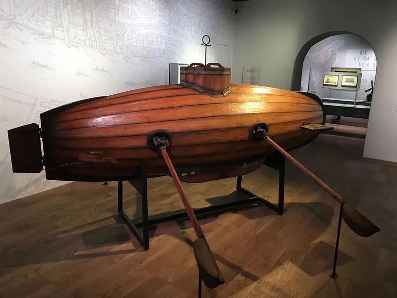 a wooden boat with two oars attached to its sides, displayed on a stand in a museum. The overlaid text reads: 'this is a…