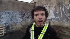 A man in a tan jacket and glasses, standing in front of a graffiti-covered wall with a metal structure behind him, looks…