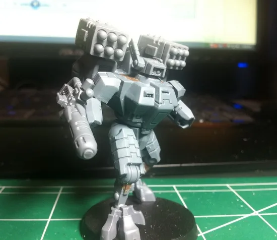 A gray, heavily armed robot miniature stands on a black base atop a green grid mat, with a computer monitor and keyboard…