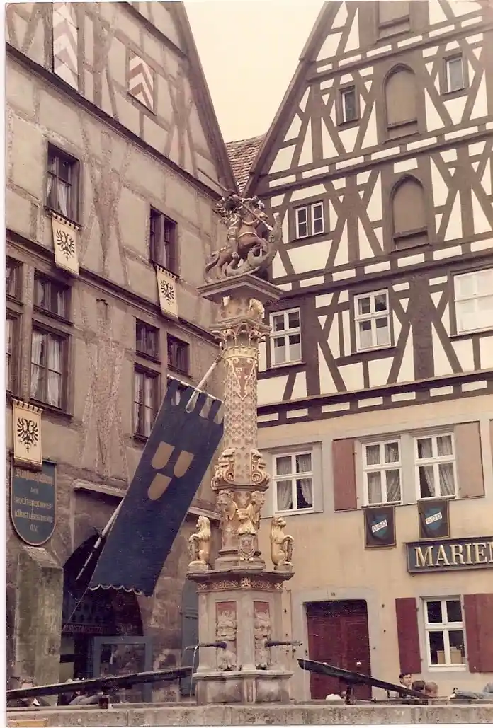 An old photograph of a European town square with half-timbered buildings and a central ornate column monument. Two cannons…