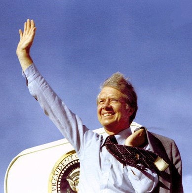A smiling man in a suit and tie waves enthusiastically while standing behind a podium with the presidential seal. His right…