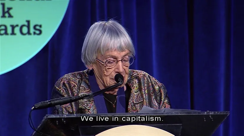 An elderly woman with gray hair and glasses stands at a podium speaking into a microphone, with a blue curtain and a…