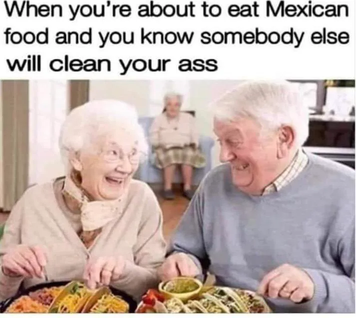An elderly couple is seated at a table, happily eating tacos and other Mexican food. The woman is laughing joyfully while…