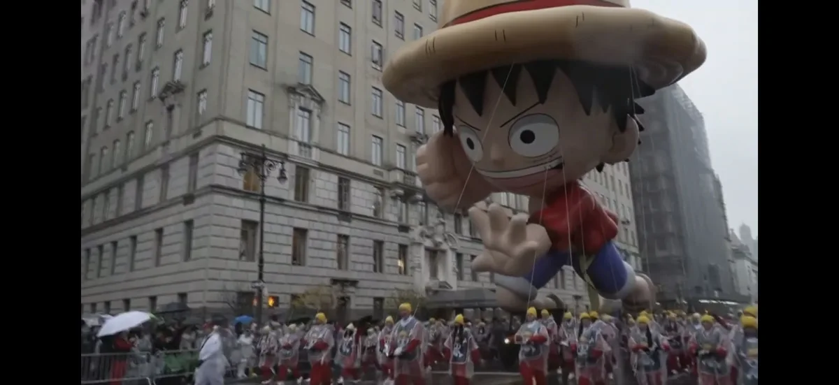 A giant inflatable balloon of the anime character Luffy from One Piece floats above a rainy city street parade, with…