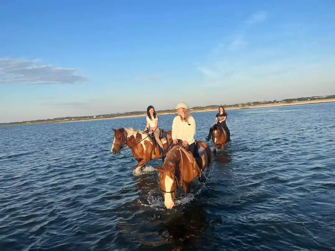 Three people are riding horses through shallow water under a clear blue sky. The central rider, wearing a light-colored…