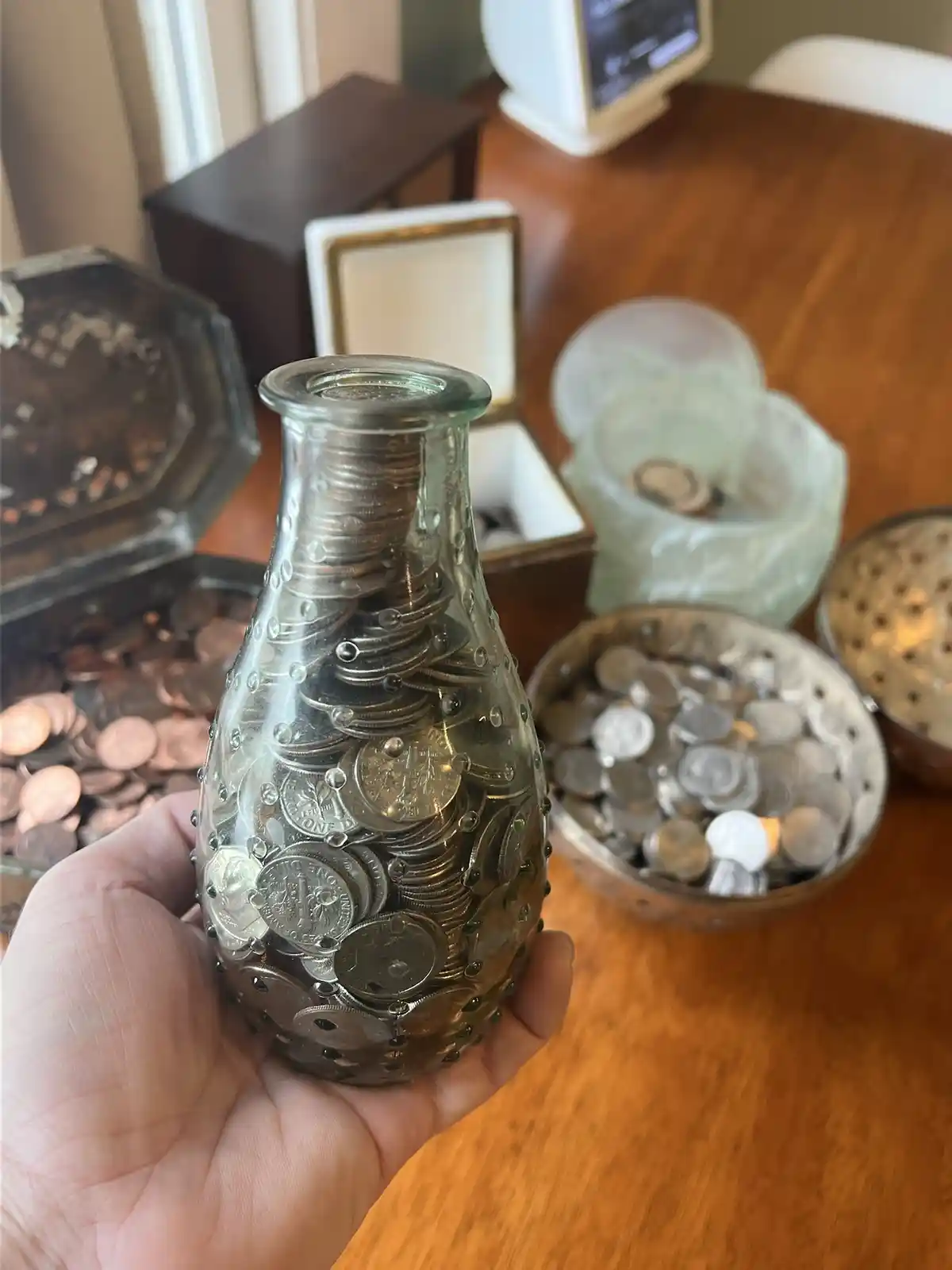 A hand holds a clear glass bottle filled with numerous silver and copper coins, with other coin-filled containers visible on…