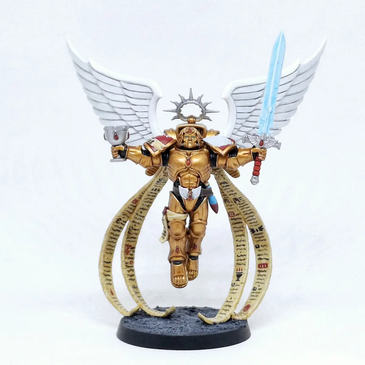 A painted miniature figurine of a muscular, winged warrior in golden armor, holding a glowing blue sword in one hand and a…