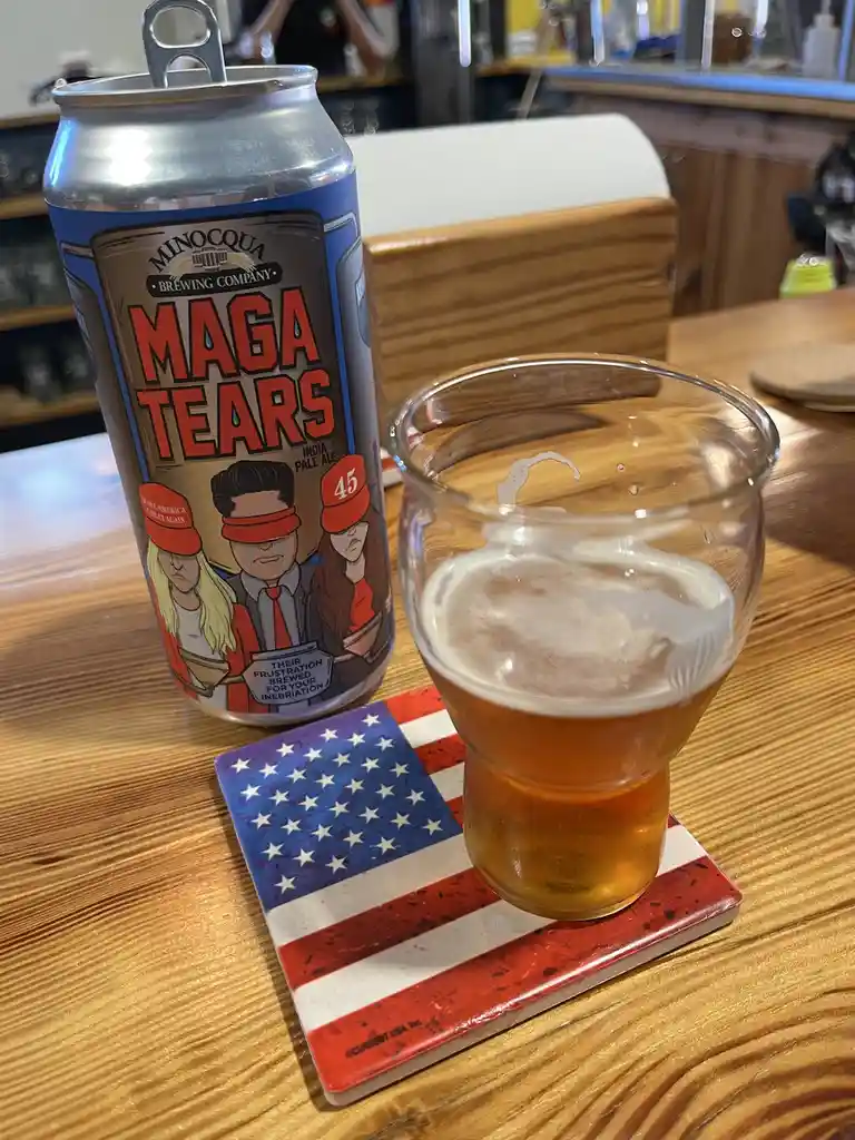 A can of Minocqua Brewing Company's 'MAGA TEARS' India Pale Ale sits next to a glass of beer on an American flag coaster…