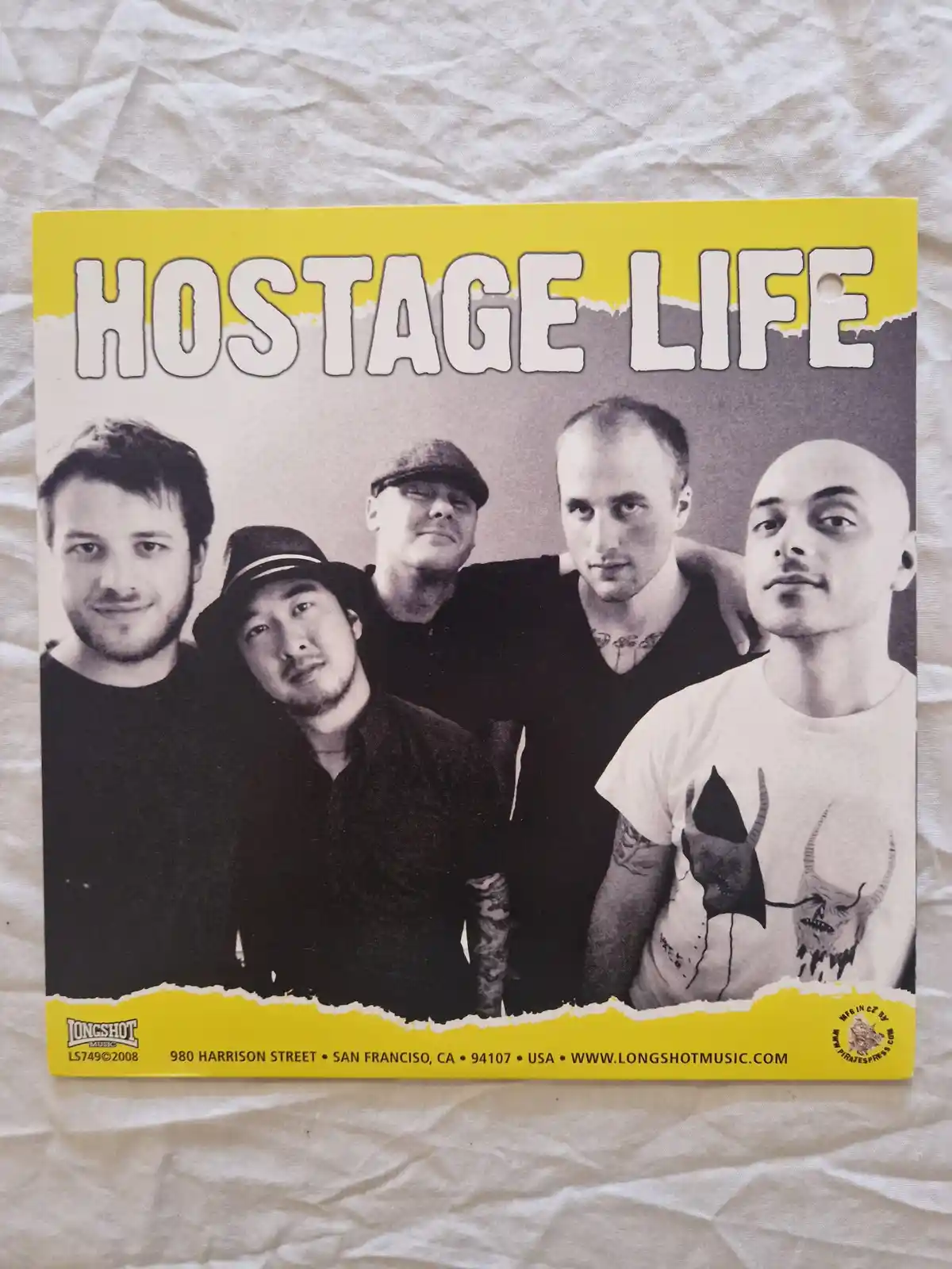 A black-and-white photo of five men, likely a band, poses on a yellow-bordered album cover titled 'HOSTAGE LIFE'. One man…