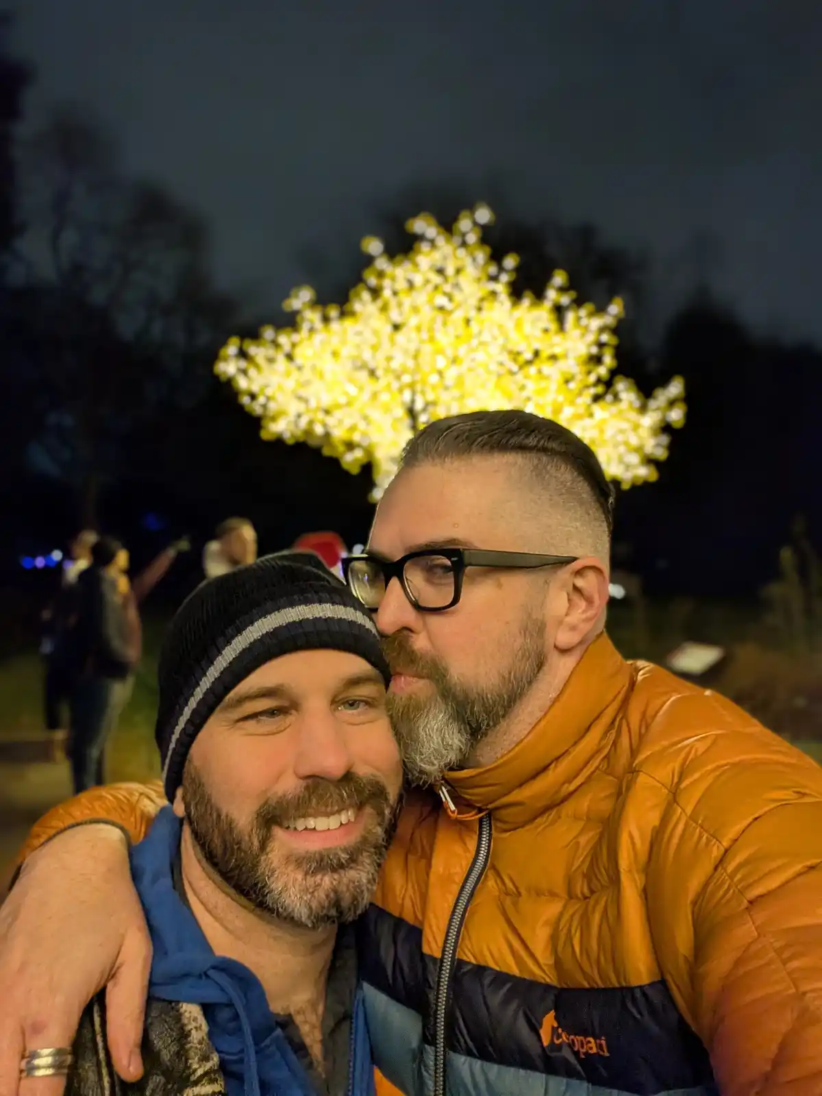 Two men are posing for a selfie at night outdoors, with one man in an orange puffer jacket and glasses leaning in close to…