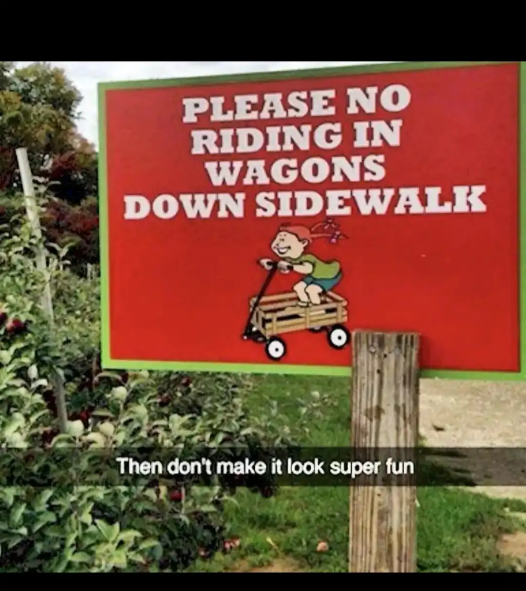 A red sign with white text reads 'PLEASE NO RIDING IN WAGONS DOWN SIDEWALK' and features a cartoon child happily riding a…