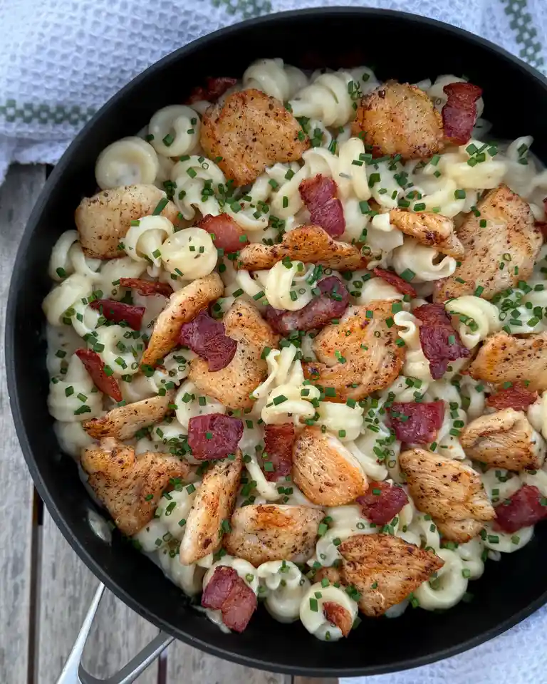 A top-down photo of a black skillet filled with creamy pasta, chunks of cooked chicken, crispy bacon pieces, and chopped…