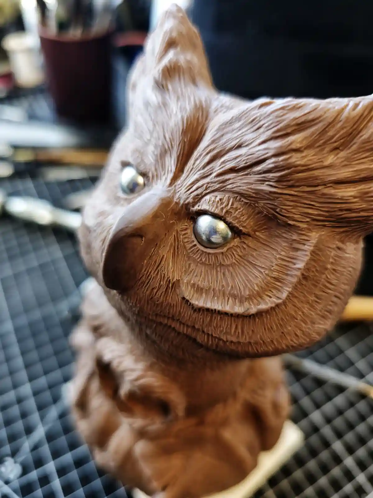 A close-up photo shows a detailed, brown clay sculpture of an owl's head with textured feathers and shiny silver eyes…