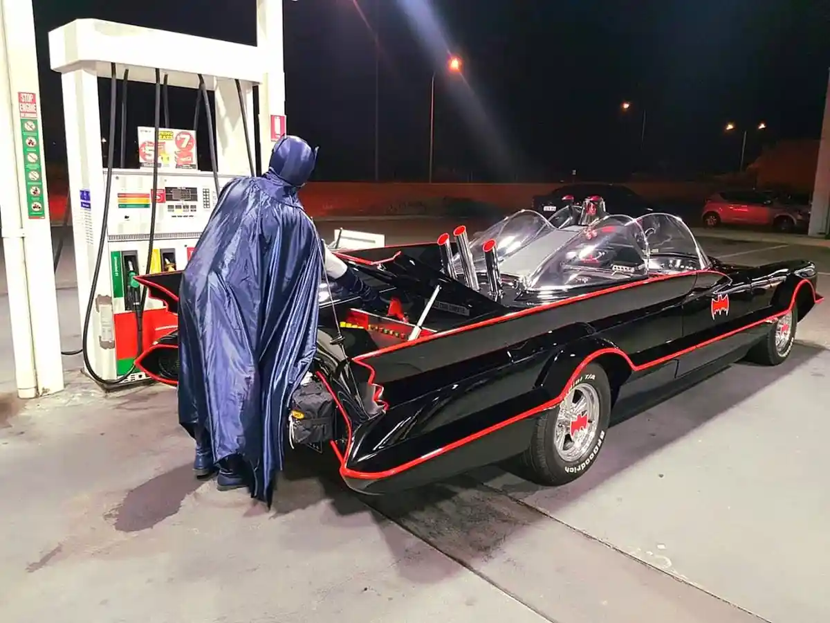 A person dressed as Batman in a blue cape and cowl is standing at a gas pump, filling up a black and red Batmobile replica…