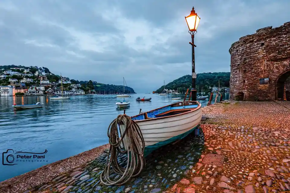 A serene harbor scene at dusk with a small white and blue rowboat resting on a cobblestone shore next to a lit vintage…