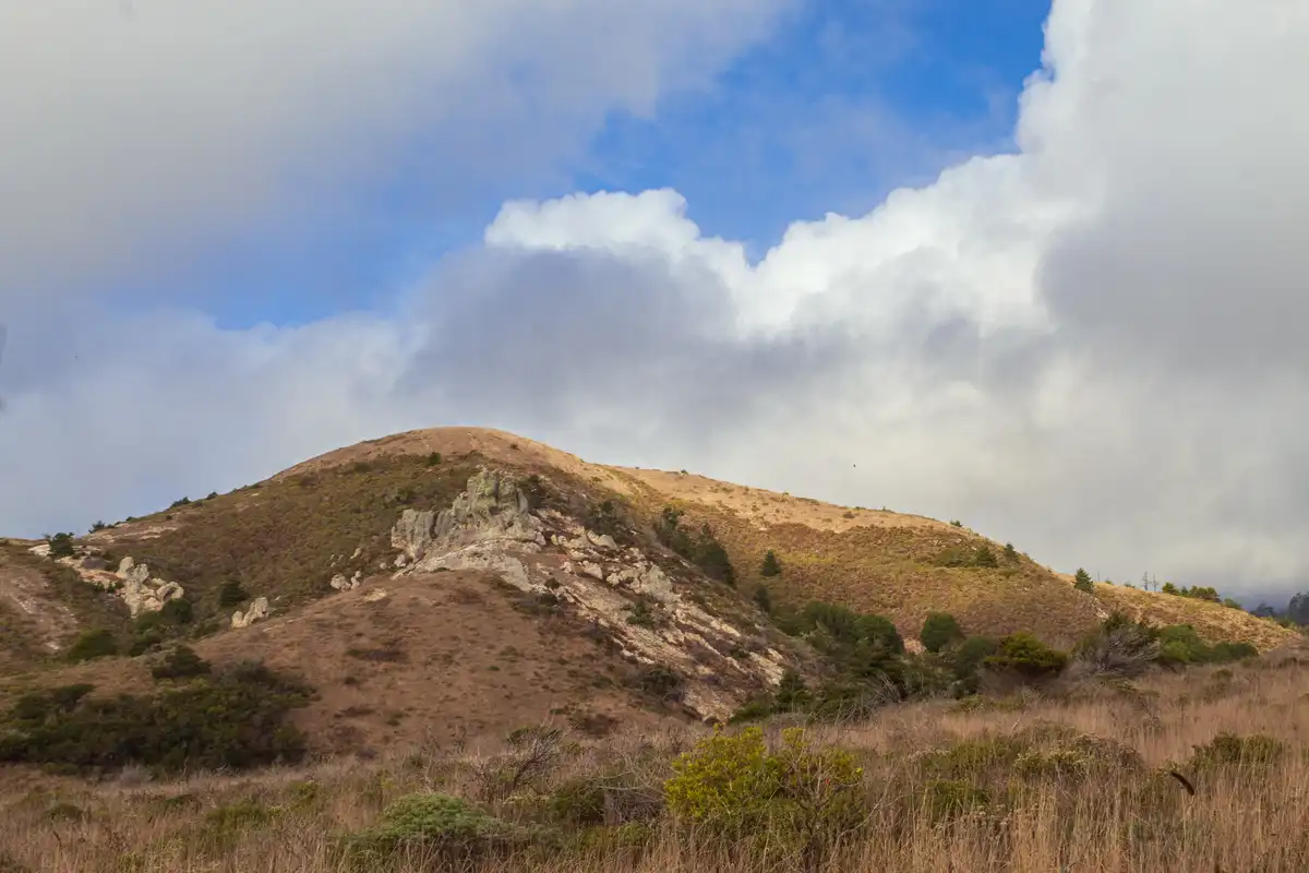 A scenic photo of a rolling, dry grass-covered hill under a partly cloudy blue sky, with scattered shrubs and rock…
